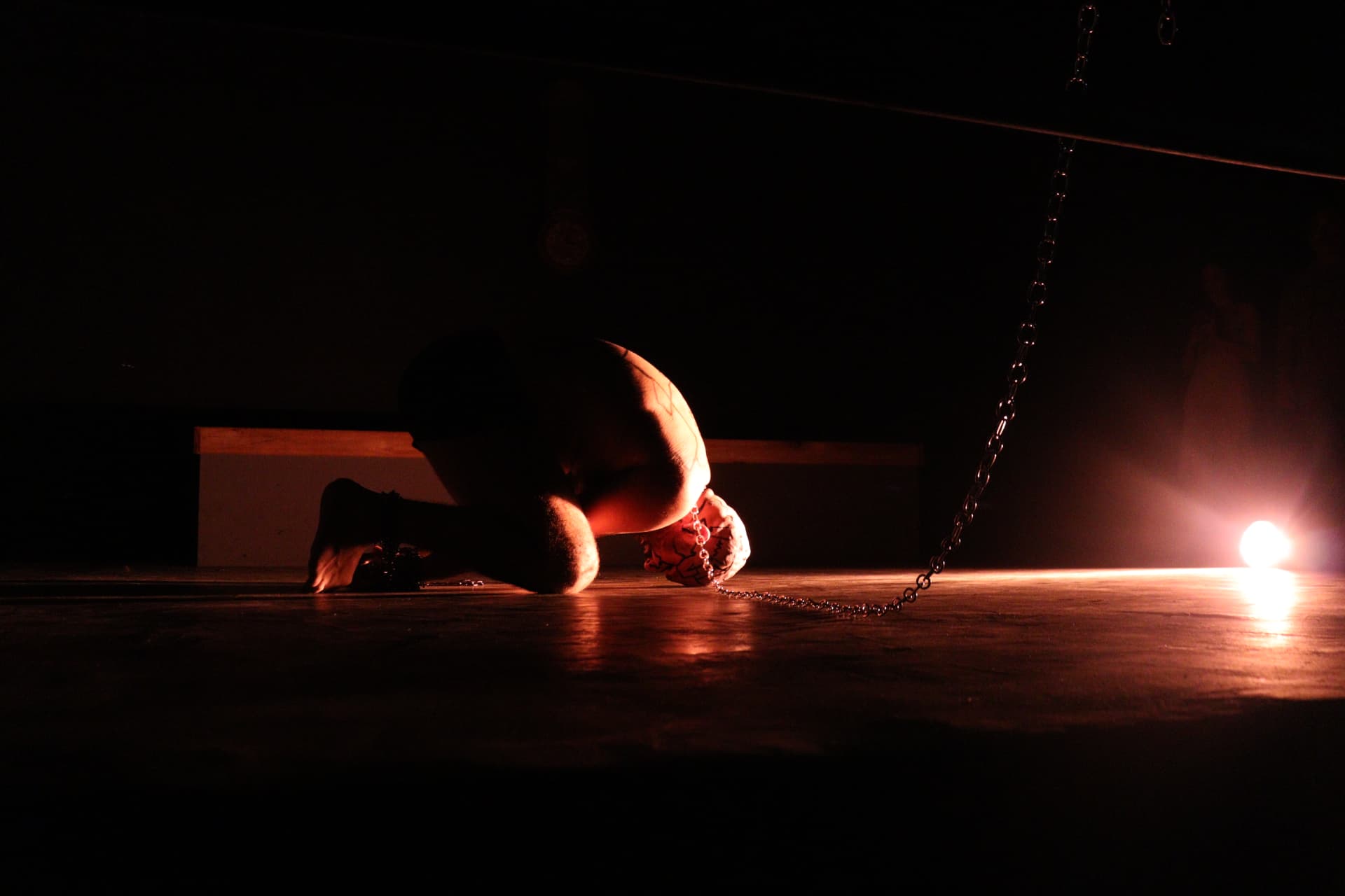 Performer curvado ao chão preso a correntes sob luz âmbar