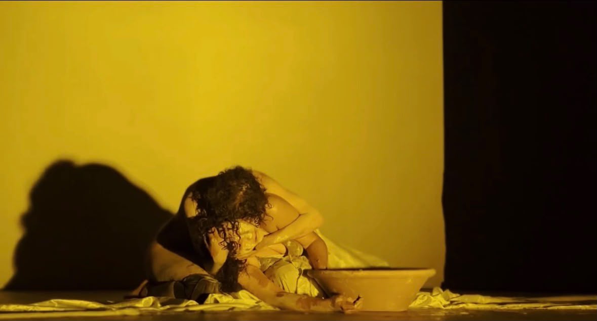 Performer curvada sobre a bacia em luz amarela, com sombra projetada na parede